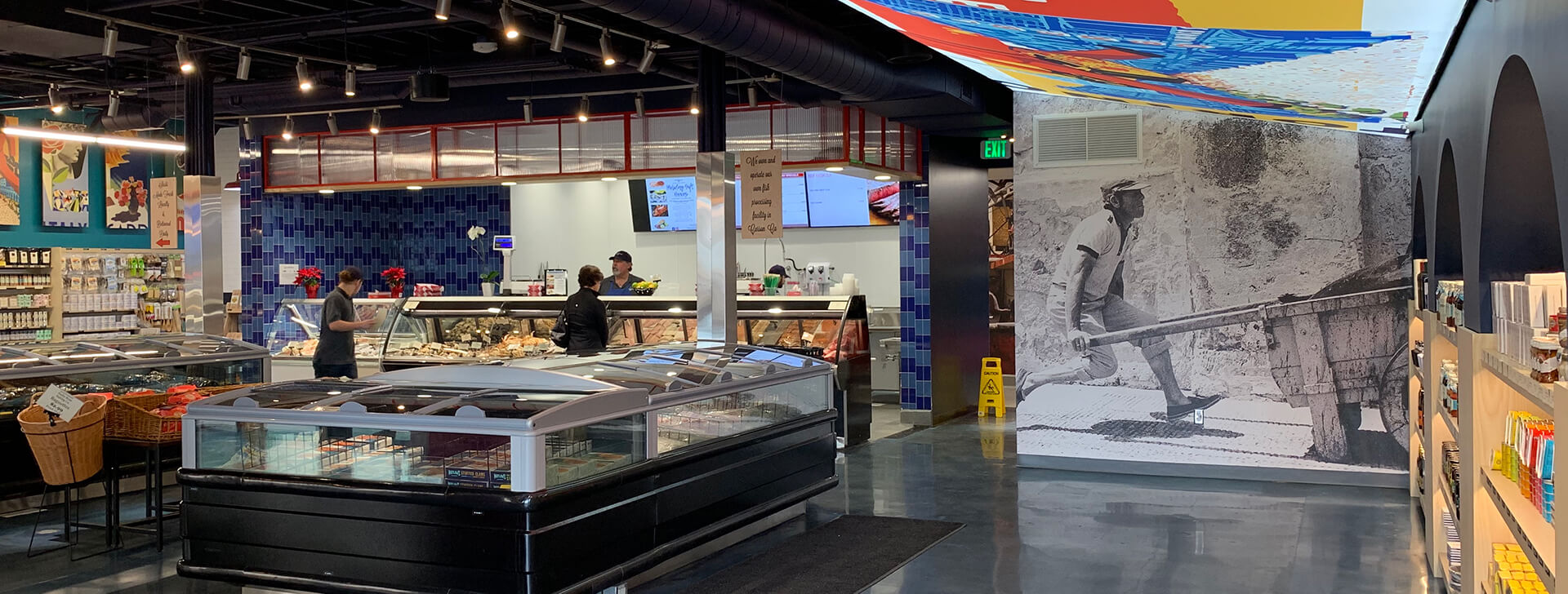 Seafood market interior with seafood cooler cases and bold interior design featuring large vibrant lighted signs, black and white photography, brightly painted walls, woven baskets and dramatic two-tone blue tiles.