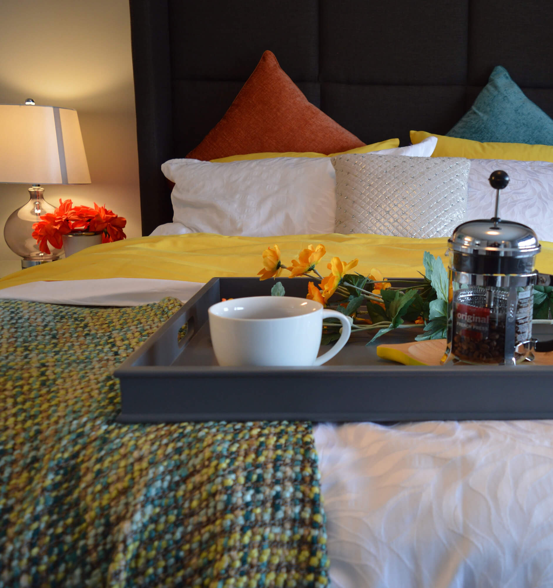 Food tray with coffe cup and carafe on end of bed in hotel guestroom decorated with colorful pillows, throw blanket, and flowers.